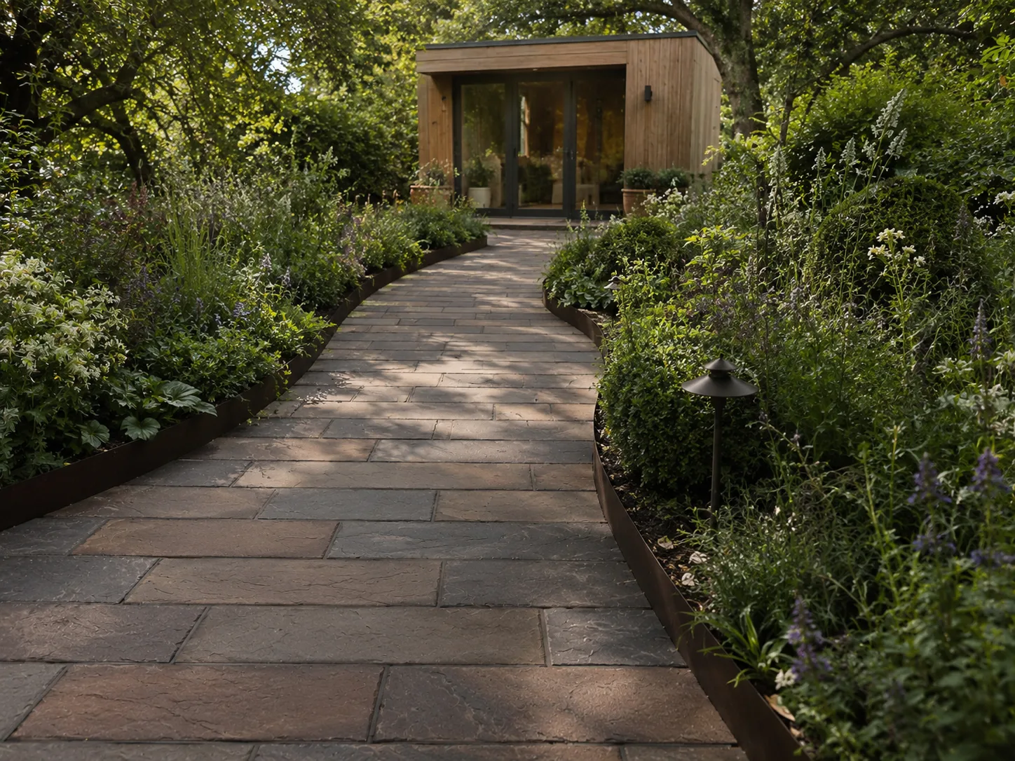 A stone path curves through a lush garden towards a wooden garden room.