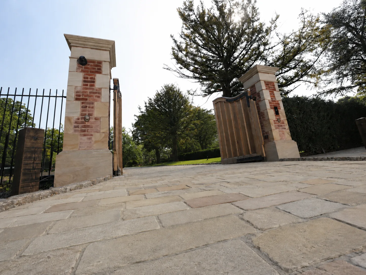 A stone driveway leads to wooden gates between two brick and stone pillars, with trees in the background.