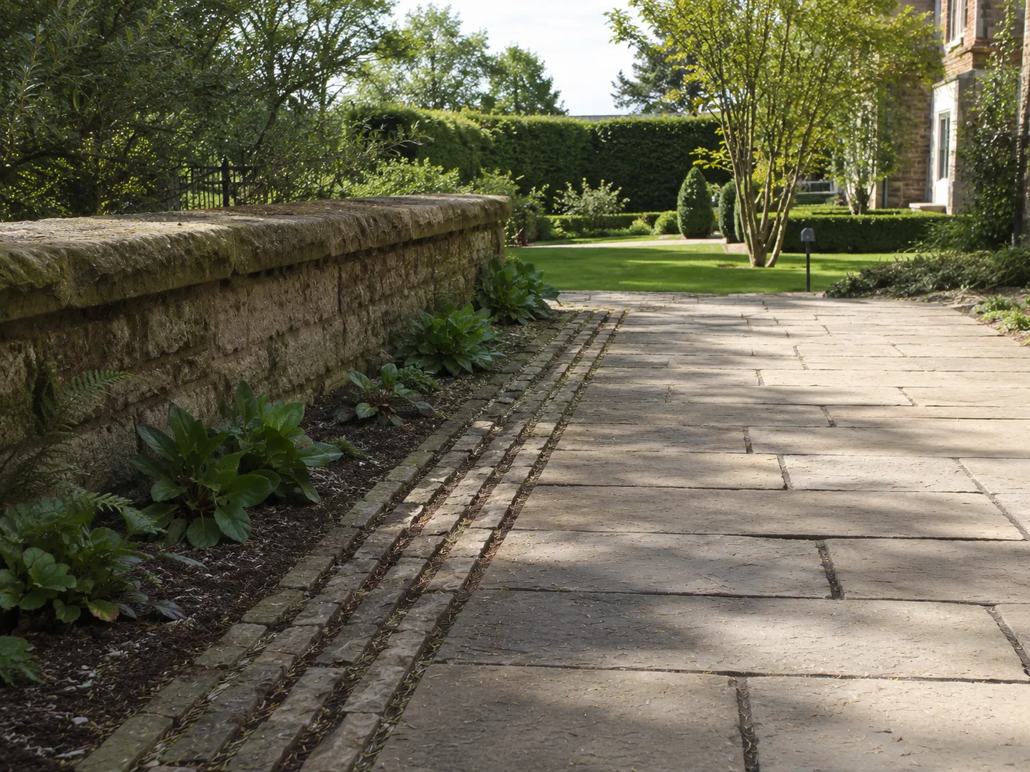 A stone path runs alongside a weathered stone wall with plants growing at its base, leading towards a garden with trees and hedges.