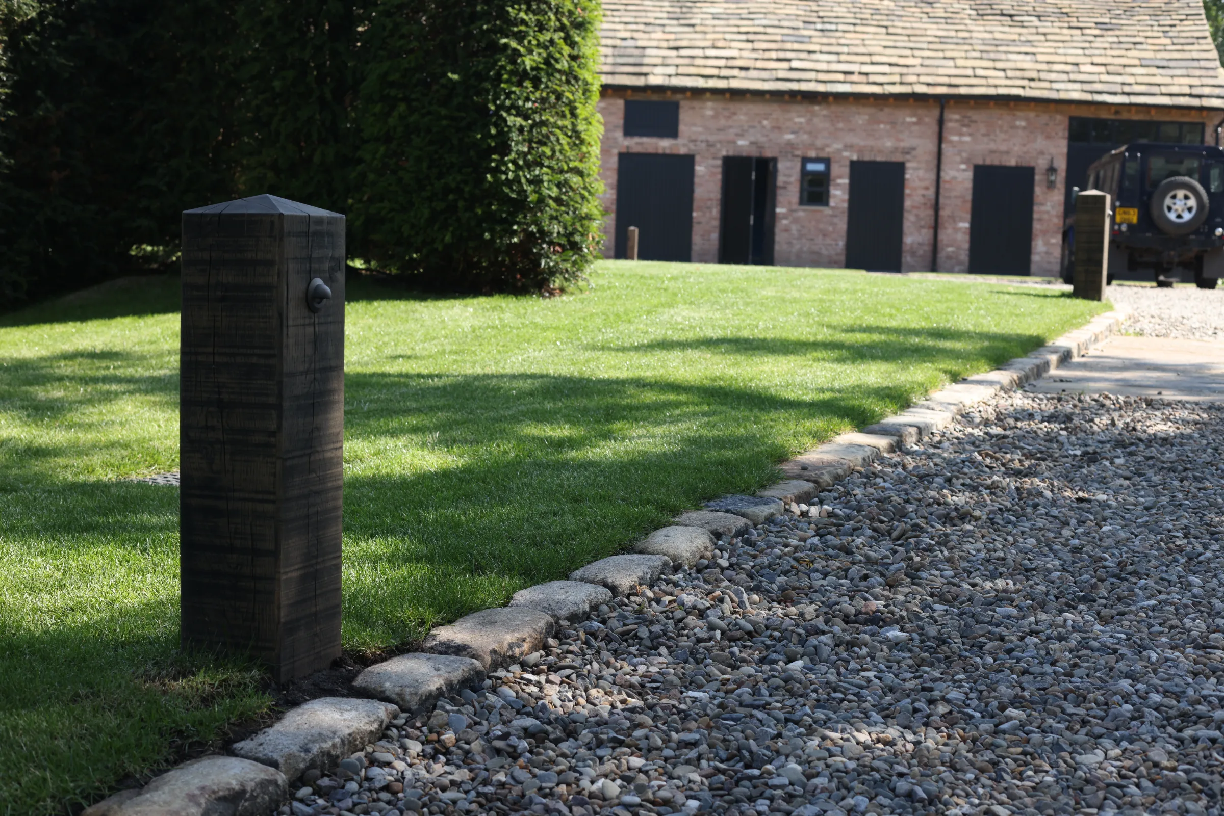 A gravel path bordered by a lawn and a wooden post, with a brick building and parked vehicle in the background.
