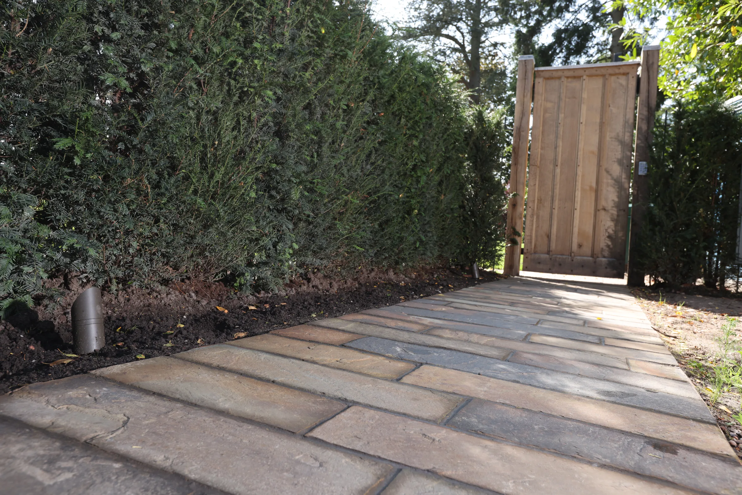 A stone path bordered by hedges leads to a wooden gate under sunlight.