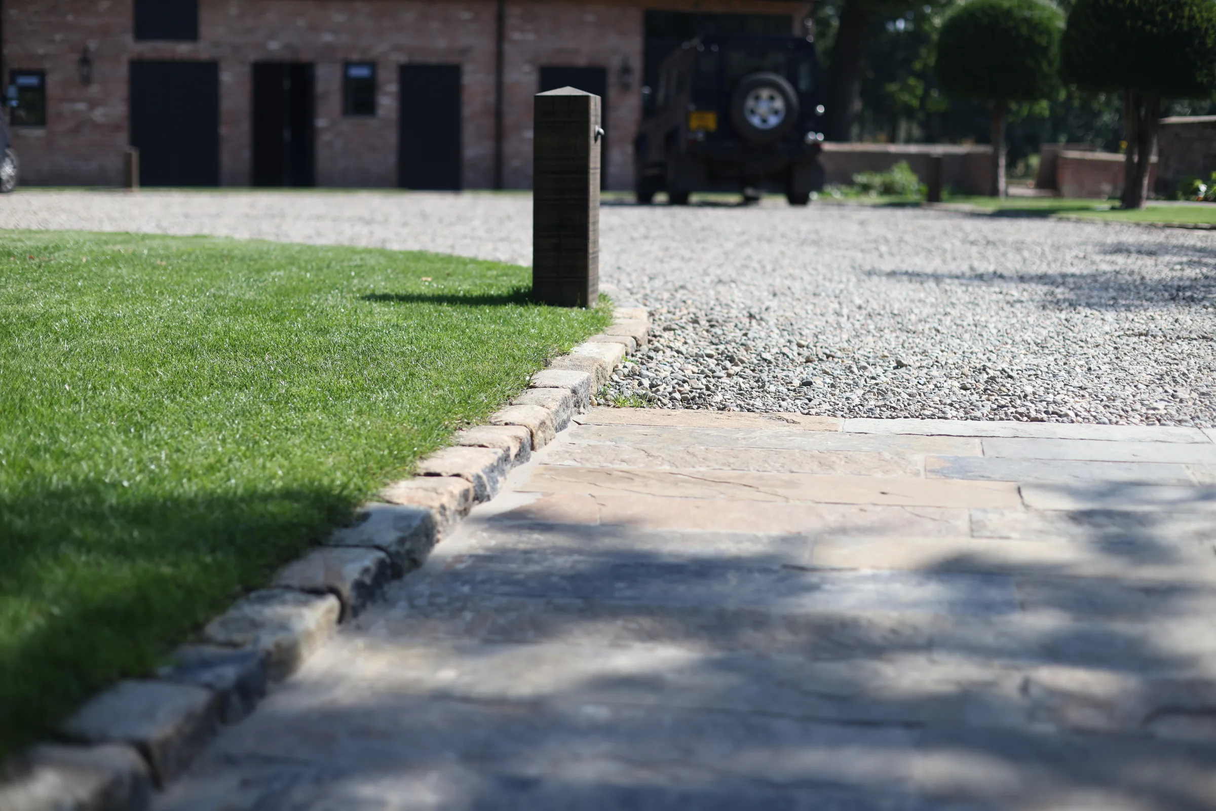 A stone path bordered by grass leads to a gravel driveway with a brick building and a vehicle in the background.