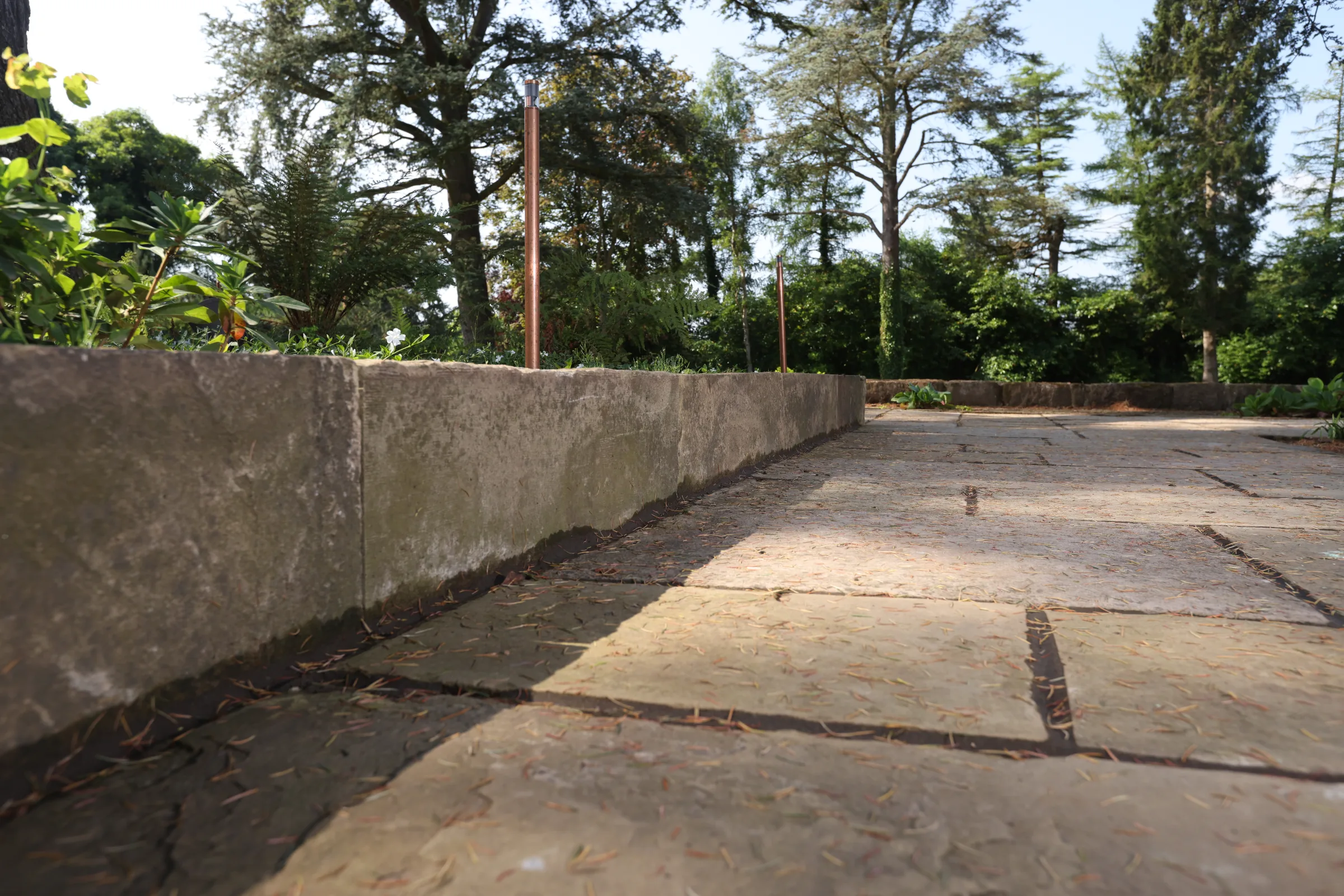 A stone terrace with large square slabs, bordered by a low stone wall, surrounded by trees and greenery.