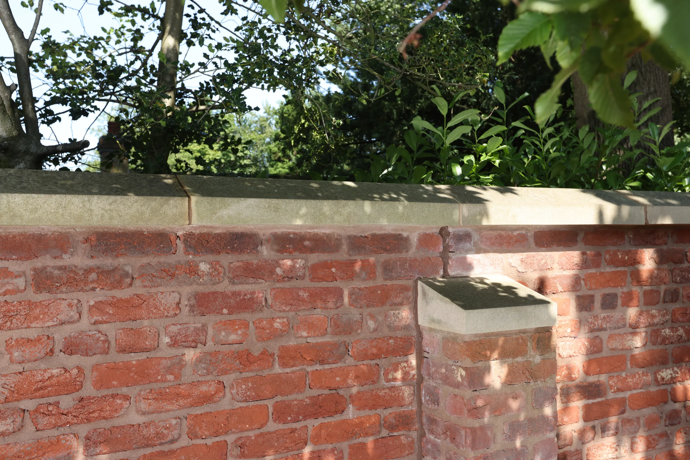 A red brick wall with a stone coping, partially in sunlight, with trees and leaves in the background.