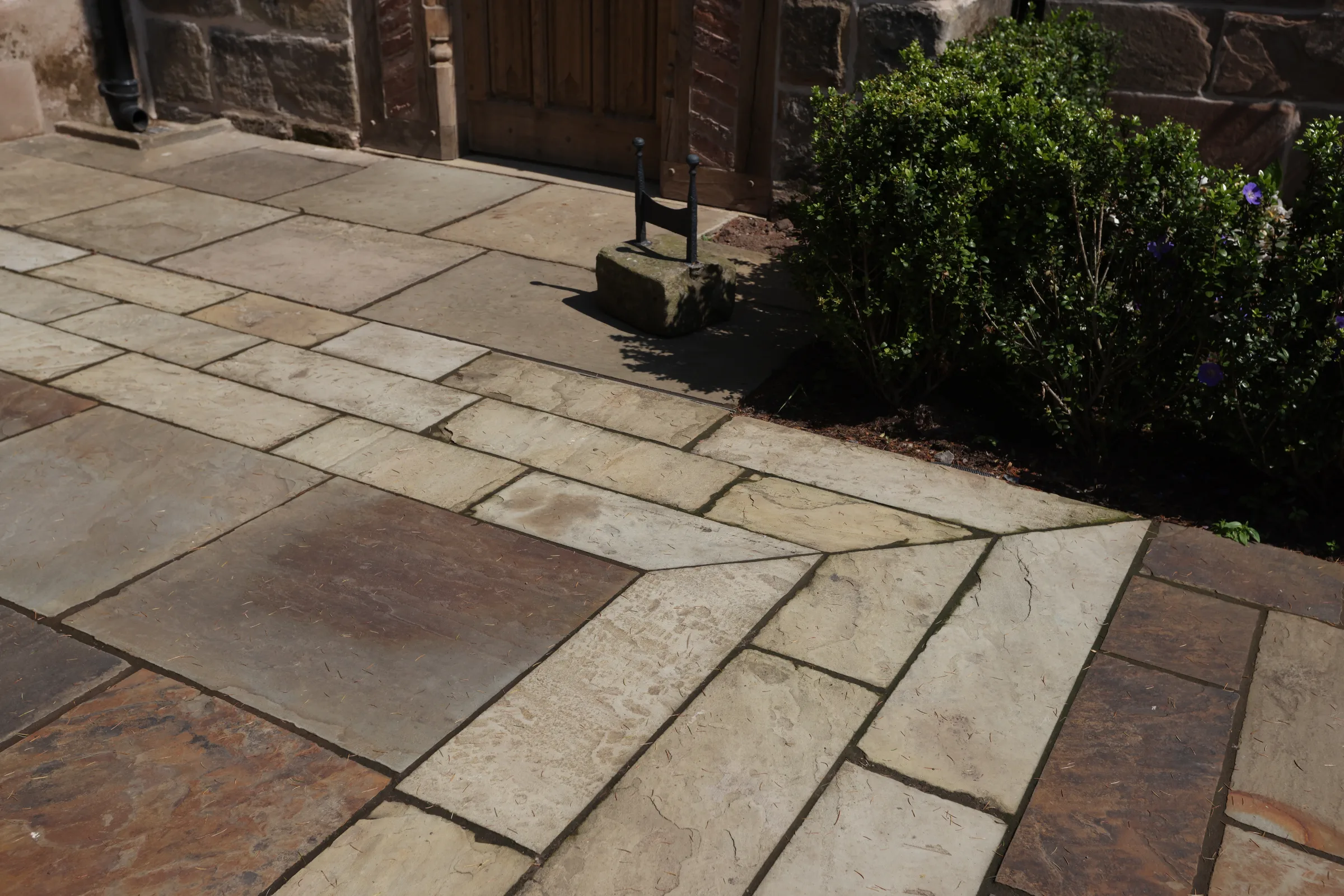 A stone patio with variously sized and coloured Yorkstone flags, bordered by a small shrub and a wooden door in the background.