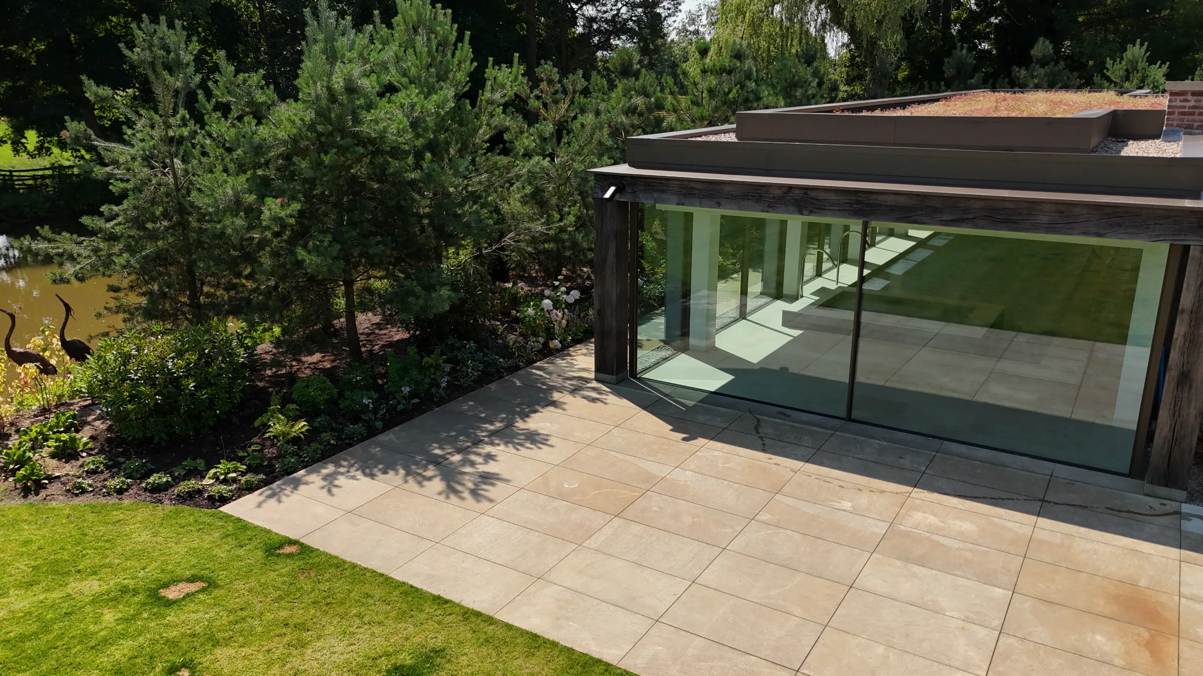 A modern glass extension with a paved terrace, surrounded by trees and grass.