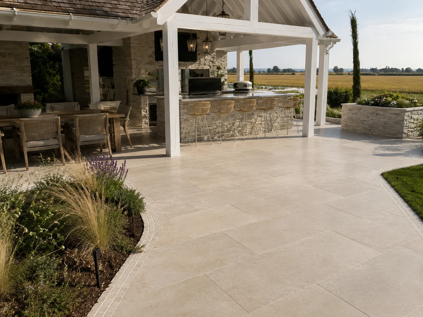 A spacious outdoor patio with pale stone tiles, a covered seating area, and a view of open fields.