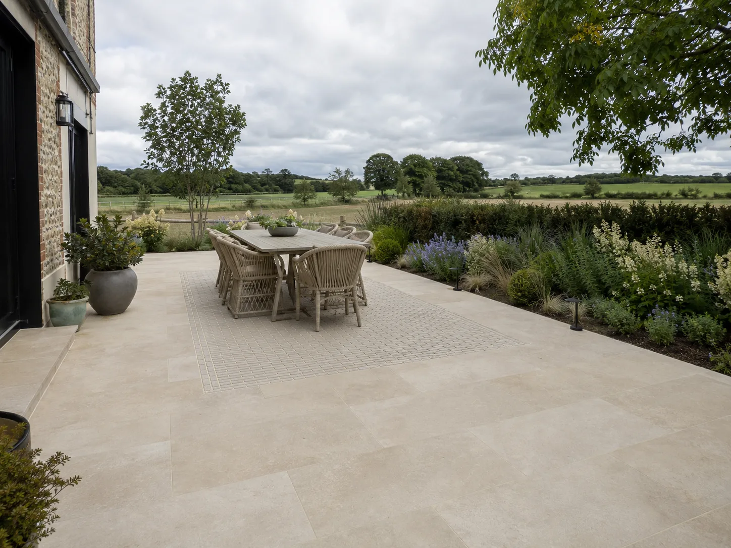A patio with pale stone tiles, a wooden dining set, and surrounding greenery, overlooking fields.
