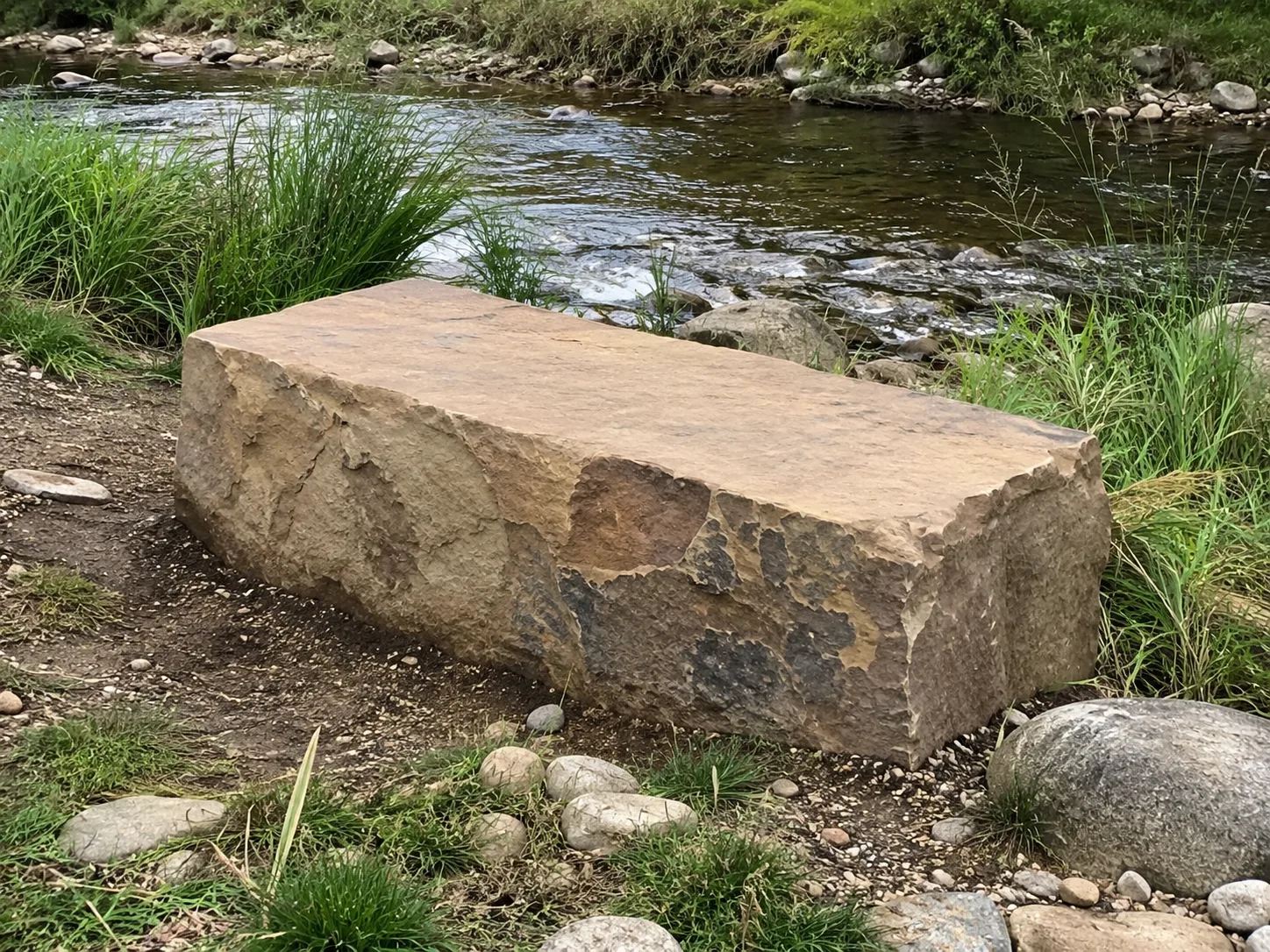 A large, rectangular stone block situated beside a flowing river, surrounded by grass and rocks.