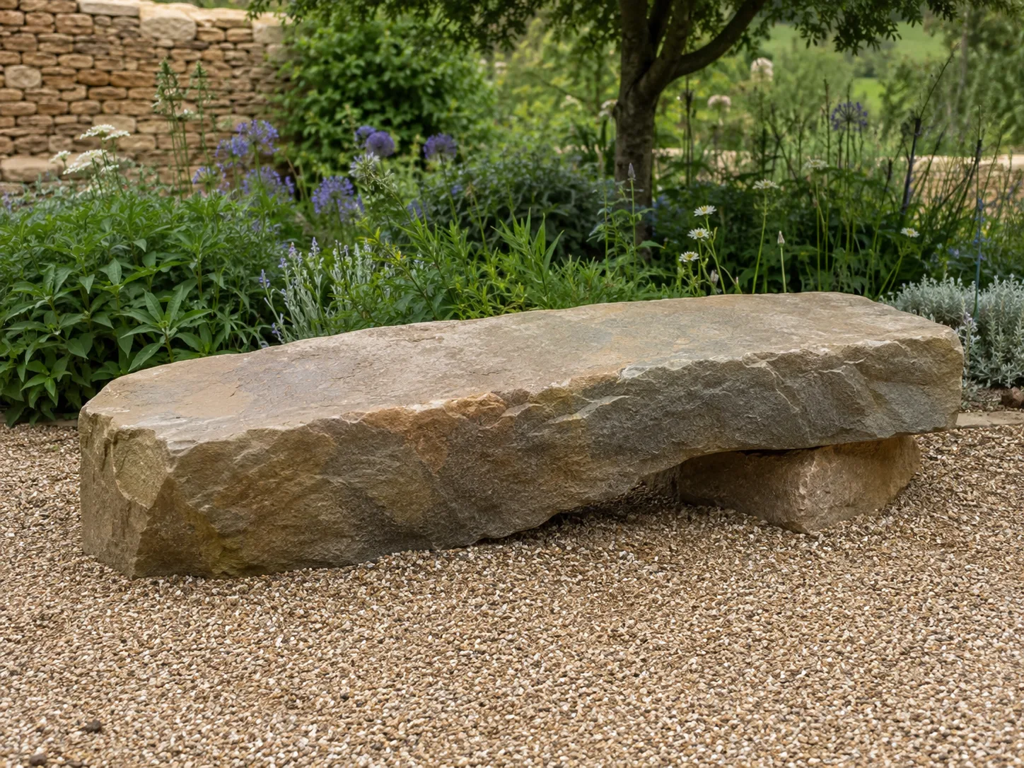 A rough-hewn stone bench set on gravel in a garden with plants and a tree in the background.