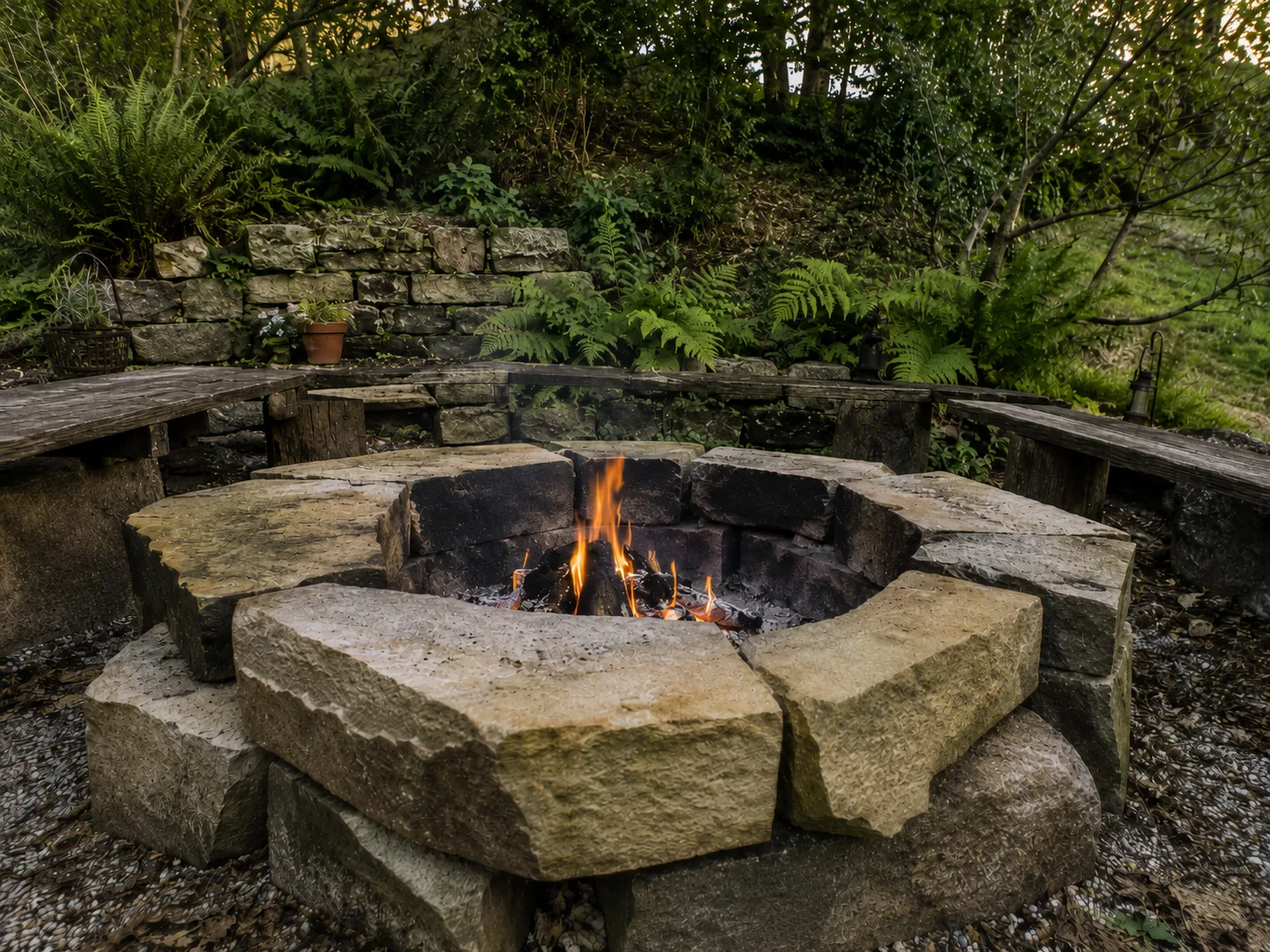 A stone fire pit with a small fire burning, surrounded by wooden benches and greenery.