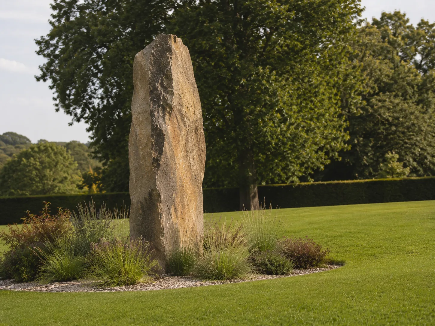 A tall, rough-hewn stone standing upright in a grassy area, surrounded by small plants and trees in the background.