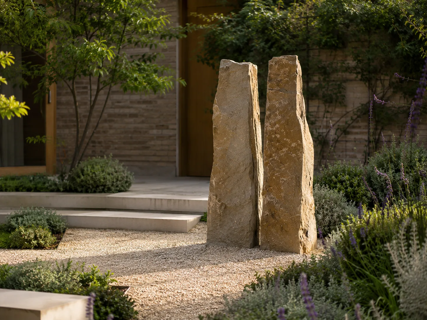 Two tall, narrow stone monoliths stand upright in a landscaped garden with gravel and greenery.