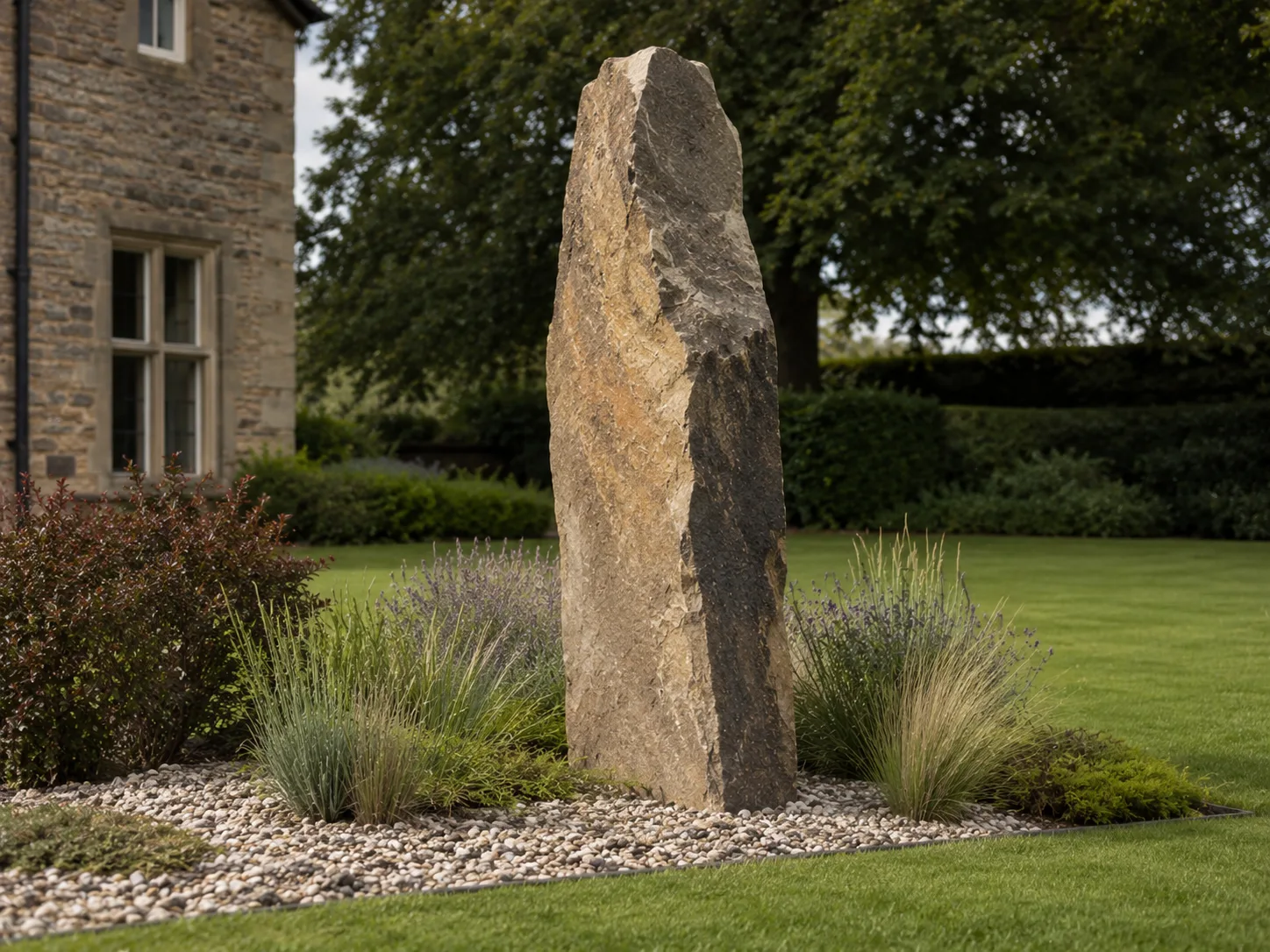 A tall, rough stone monolith set in a landscaped garden with grass, shrubs, and a stone house in the background.