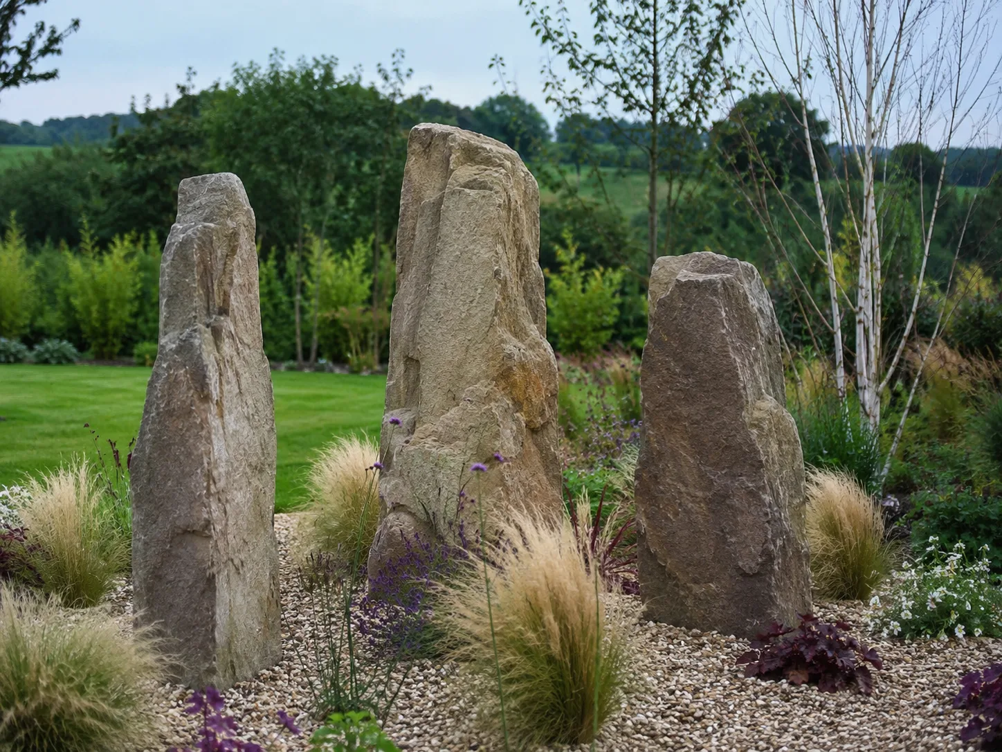 Three upright stone monoliths surrounded by gravel and plants in a garden setting.