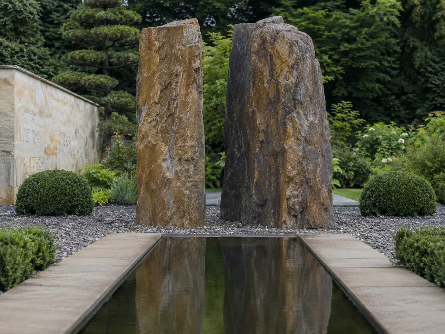 Two large standing stones beside a reflective water feature in a landscaped garden with trimmed bushes and trees.