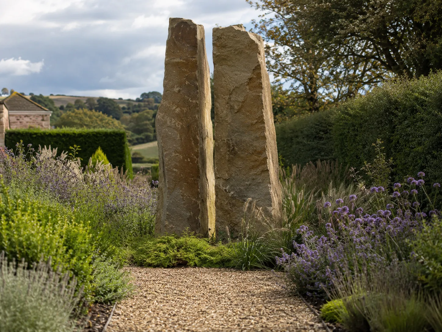 Two tall standing stones in a landscaped garden with gravel path and surrounding plants.