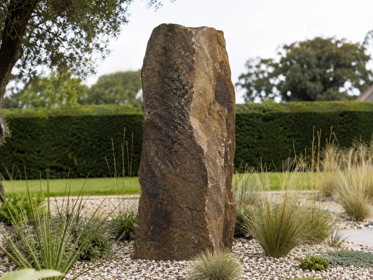 A tall, rough-hewn stone standing upright in a landscaped garden with gravel and ornamental grasses.