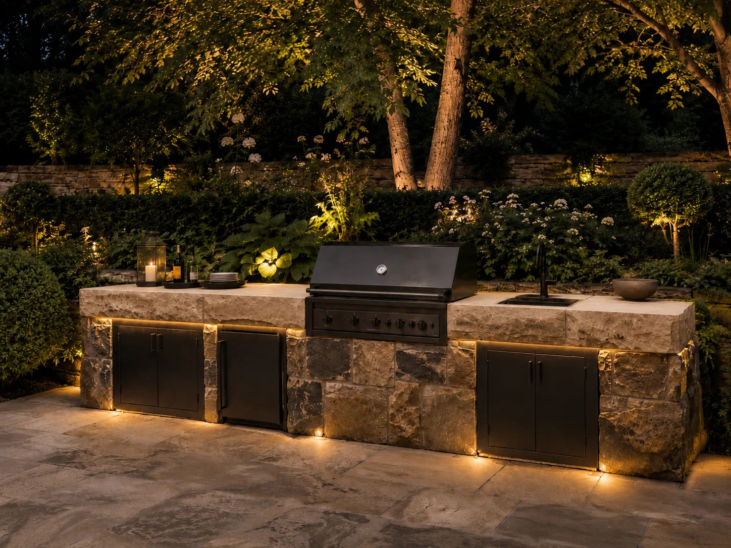 An outdoor kitchen with a stone countertop and built-in grill, surrounded by illuminated plants and trees at dusk.